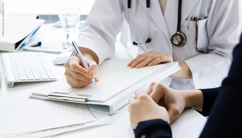 Foto mit Blick auf die Hände einer Ärztin und eines Patienten in einer Gesprächssituation an einem Tisch, die Ärztin füllt ein Formular aus